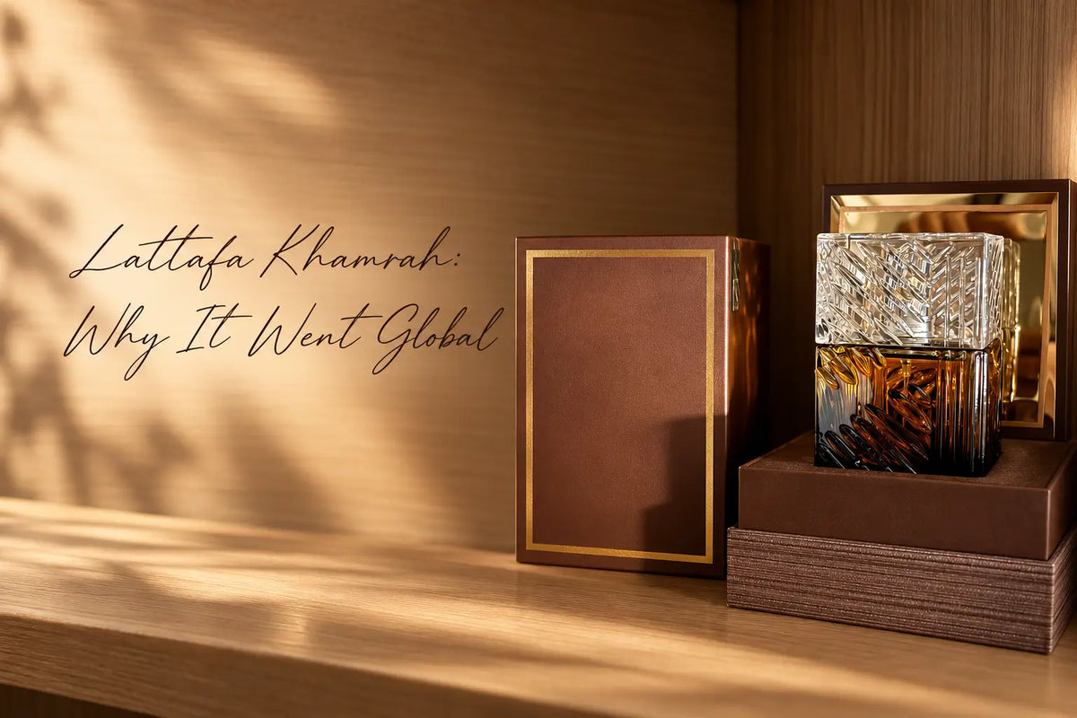 Faceted amber and clear glass perfume bottle in an open brown and gold presentation box beside a closed brown box on a wooden shelf in dappled sunlight, with the words Lattafa Khamrah Why It Went Global in script