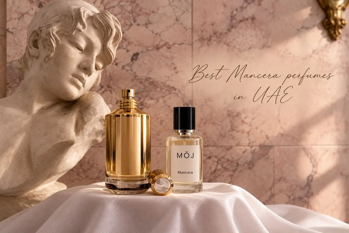 MOJ Mancera bottle beside a tall gold Mancera bottle with its cap off on a white cloth, a marble bust and pink marble wall behind, Best Mancera Perfumes in UAE
