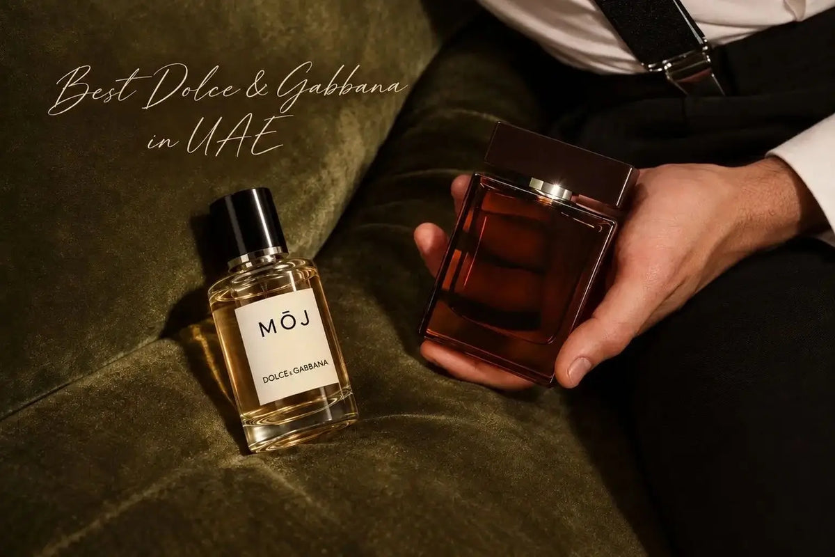 MOJ perfume bottle labelled Dolce & Gabbana on a green velvet sofa beside a dark amber Dolce & Gabbana bottle held in a man’s hand