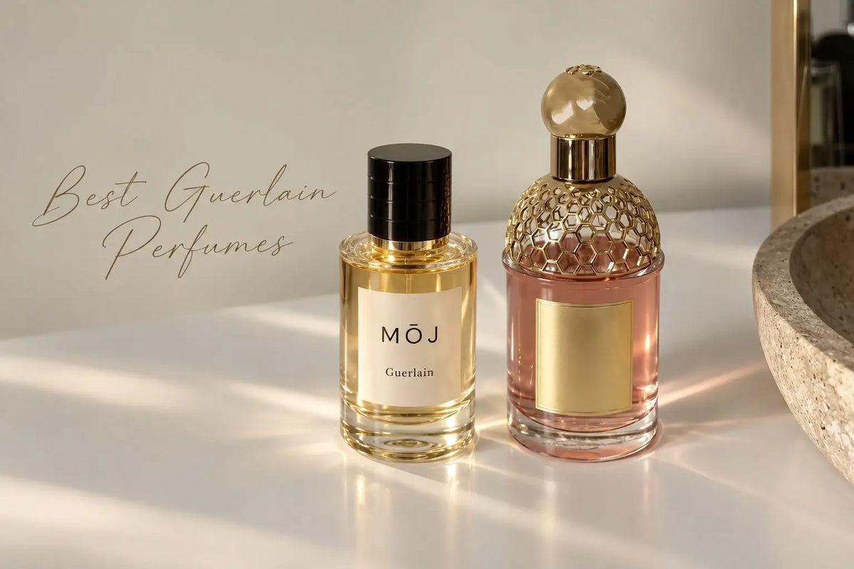 MOJ Guerlain perfume bottle with black cap beside a Guerlain bee bottle with a gold honeycomb cap and pink juice, both on a white marble counter in soft sunlight