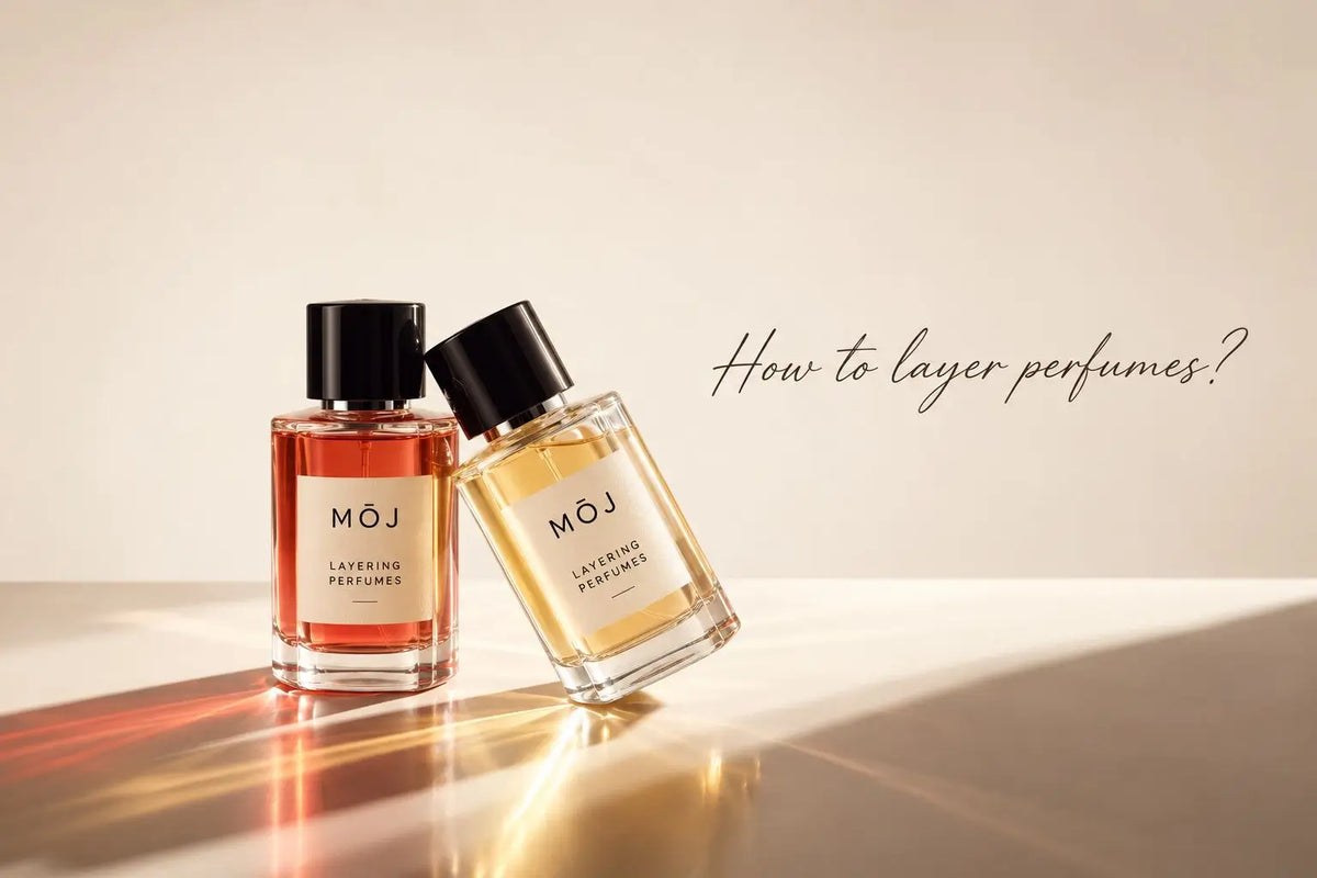 Two MOJ Layering Perfumes bottles, one amber red and one pale gold, leaning together in sunlight on a cream surface, How to Layer Your Perfumes in the UAE